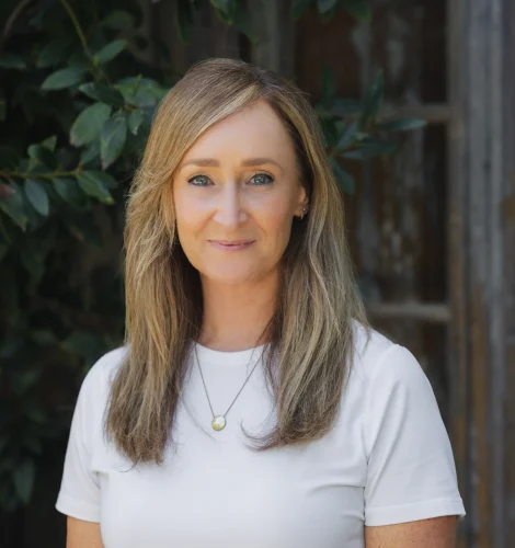 Close-up portrait of a woman with shoulder-length light brown hair wearing a white shirt, standing outdoors with green leaves in the background.