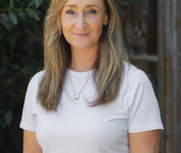 Close-up portrait of a woman with shoulder-length light brown hair wearing a white shirt, standing outdoors with green leaves in the background.