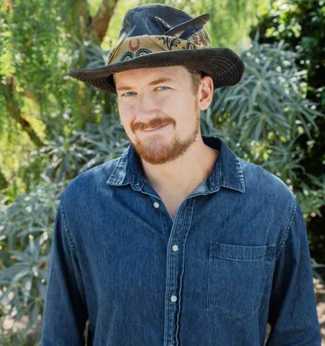 Portrait of a man wearing a denim shirt and a wide-brimmed hat, standing in a lush garden backdrop.
