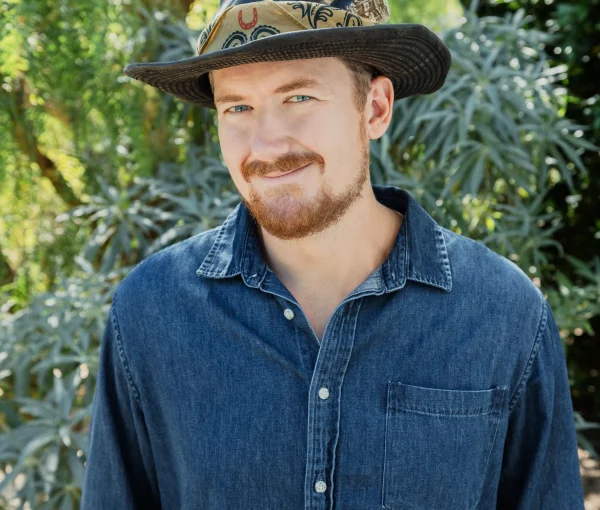 Portrait of a man wearing a denim shirt and a wide-brimmed hat, standing in a lush garden backdrop.