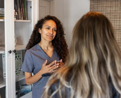 Female healthcare professional in blue scrubs converses with a patient in a room with shelves of medical books behind them.