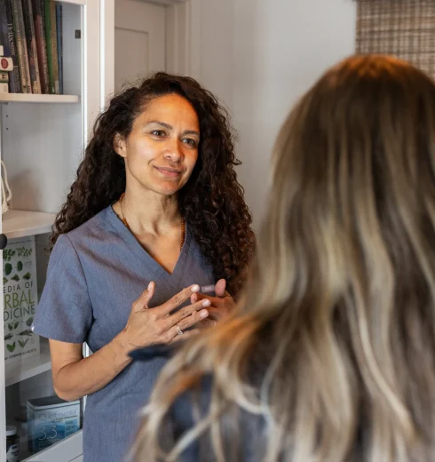 Female healthcare professional in blue scrubs converses with a patient in a room with shelves of medical books behind them.