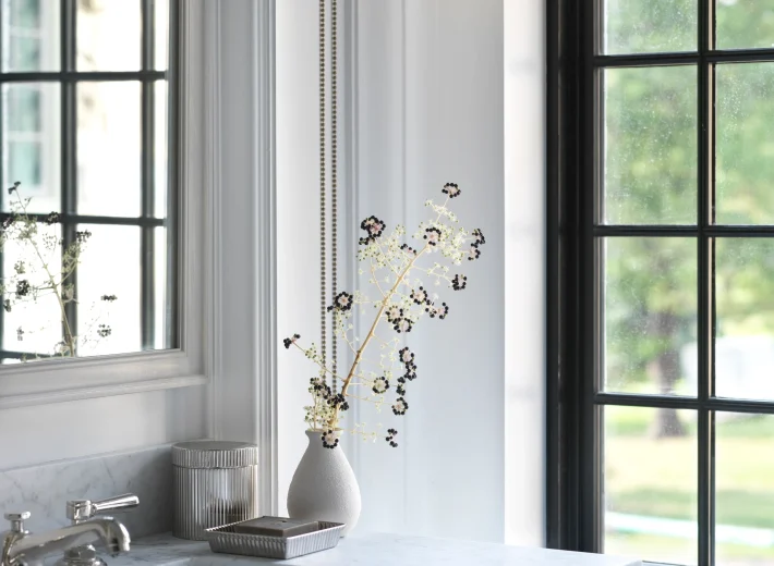 White kitchen counter with marble surface, a vase of dried flowers, and a window with beige roman blinds nearby.