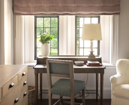 Wood desk and chair by a window with a pink fabric valance, a white lamp, and a potted plant on the sill; a dark wooden dresser to the left.