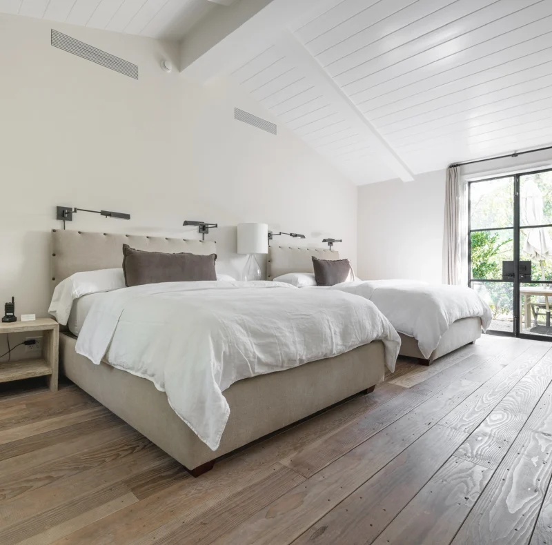 Bright hotel bedroom with two twin beds, beige headboards, wall lamps, and a balcony door with curtains to the outside.