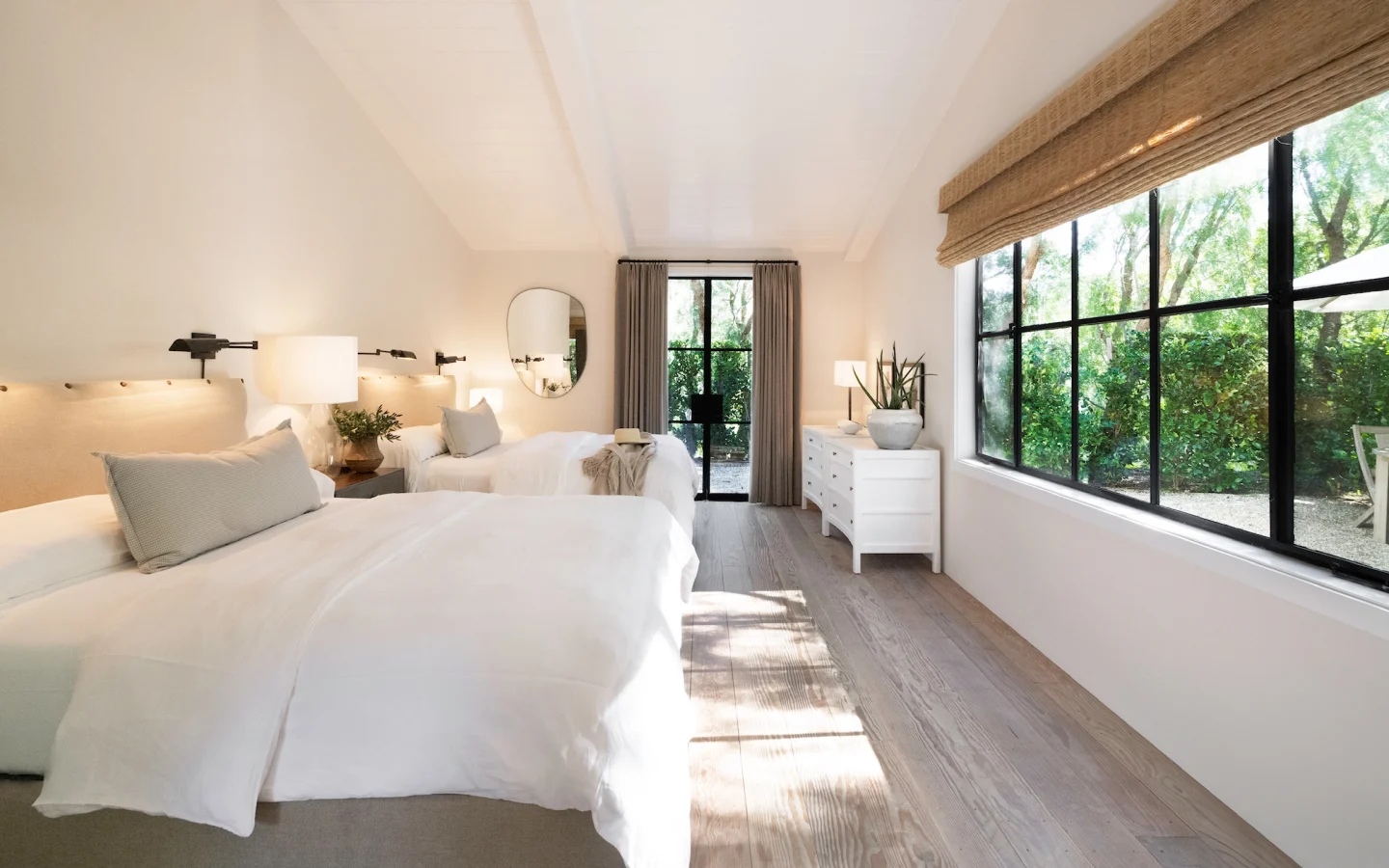 Bright, airy twin bedroom with white linens, beige headboards, wall lamps, and a large window wall overlooking greenery outside.