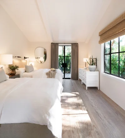 Bright, airy twin bedroom with white linens, beige headboards, wall lamps, and a large window wall overlooking greenery outside.