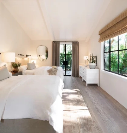 Bright, airy twin bedroom with white linens, beige headboards, wall lamps, and a large window wall overlooking greenery outside.
