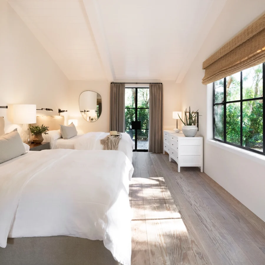 Bright, airy twin bedroom with white linens, beige headboards, wall lamps, and a large window wall overlooking greenery outside.