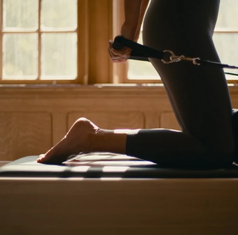 Person in black athletic wear kneels on a Pilates reformer, foot strapped to a bar during a leg extension exercise by a sunlit windowed wall.