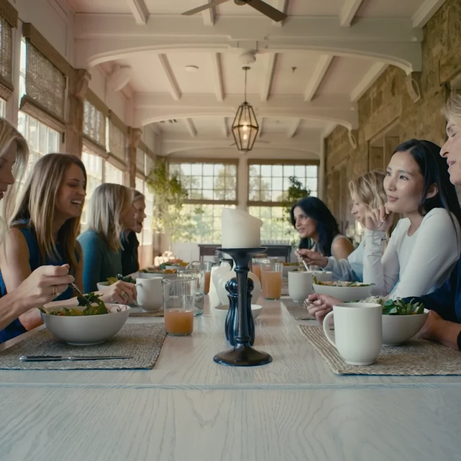 Group of people dining together at a long table in a bright stone-walled room, eating salads and chatting.