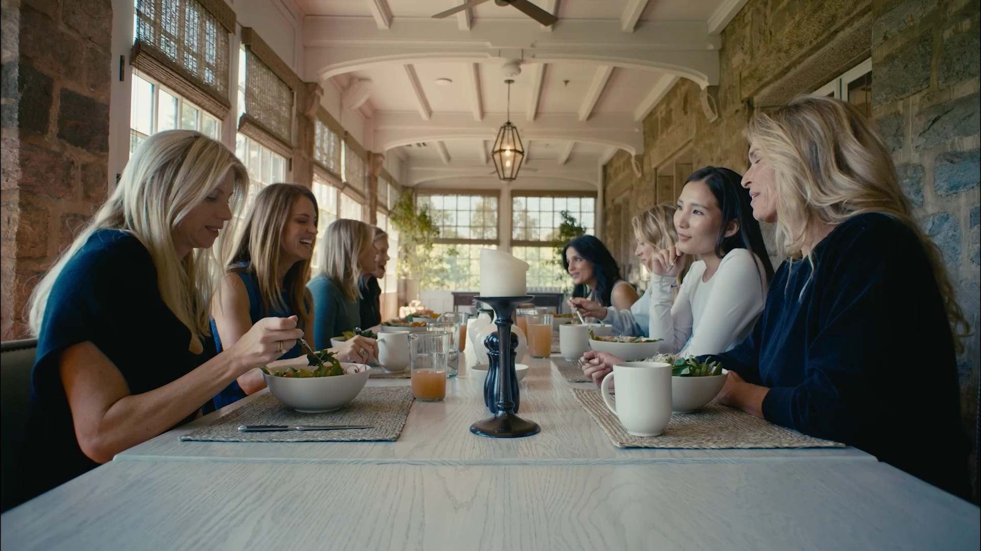 Group of people dining together at a long table in a bright stone-walled room, eating salads and chatting.