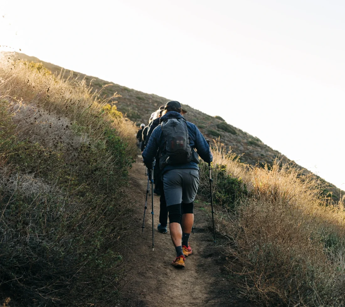 Group of hikers with backpacks walking uphill on a sunlit dirt trail, using trekking poles.