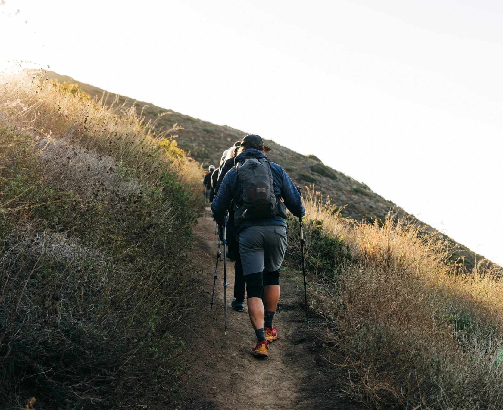 Group of hikers with backpacks walking uphill on a sunlit dirt trail, using trekking poles.