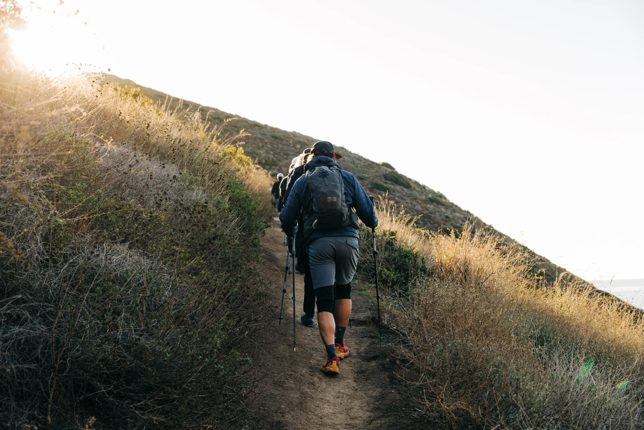Group of hikers with backpacks walking uphill on a sunlit dirt trail, using trekking poles.
