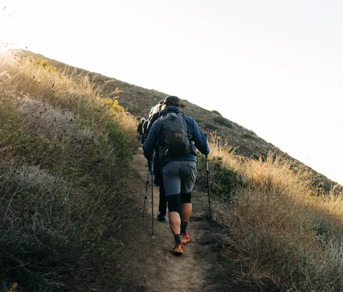 Group of hikers with backpacks walking uphill on a sunlit dirt trail, using trekking poles.