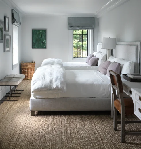 Two twin beds with white linens and gray upholstered headboards in a bright gray bedroom with a large window and gray Roman shades.