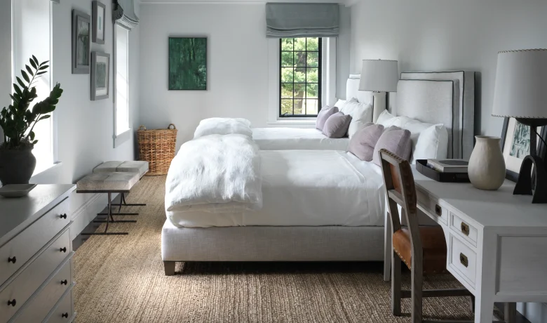 Two twin beds with white linens and gray upholstered headboards in a bright gray bedroom with a large window and gray Roman shades.