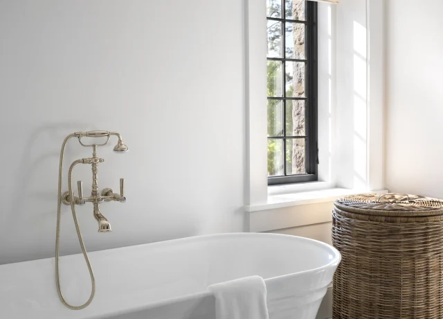 White freestanding bathtub with a brass faucet, towel draped over the rim, near a woven wicker laundry basket and a black-and-white checkered floor by a window.
