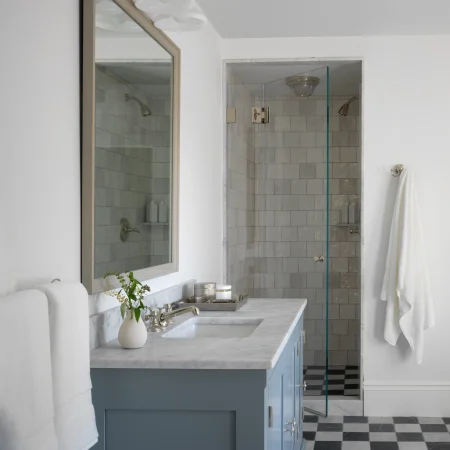 Blue vanity with white marble countertop in a modern bathroom, mirror above, towel on rack, and a glass-doored shower beyond.
