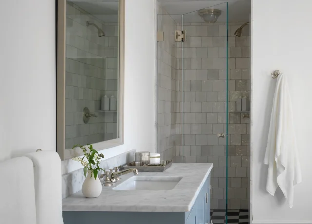Blue vanity with white marble countertop in a modern bathroom, mirror above, towel on rack, and a glass-doored shower beyond.