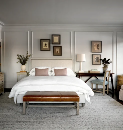 Light gray bedroom with paneled walls, white bed, and framed art above; brown leather bench at foot of bed.
