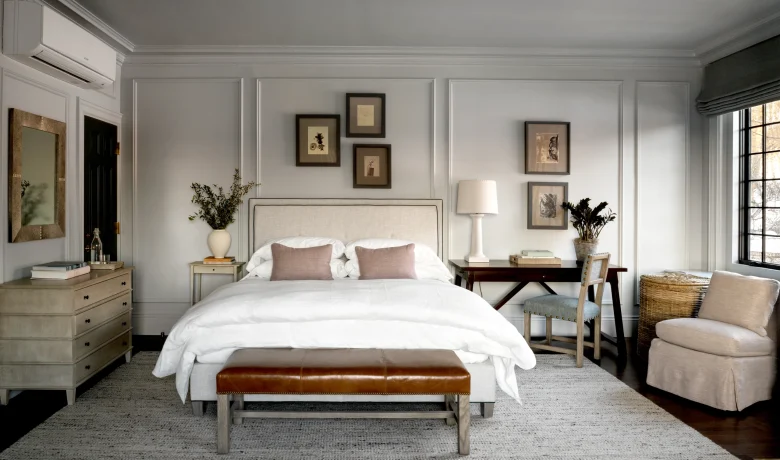 Light gray bedroom with paneled walls, white bed, and framed art above; brown leather bench at foot of bed.