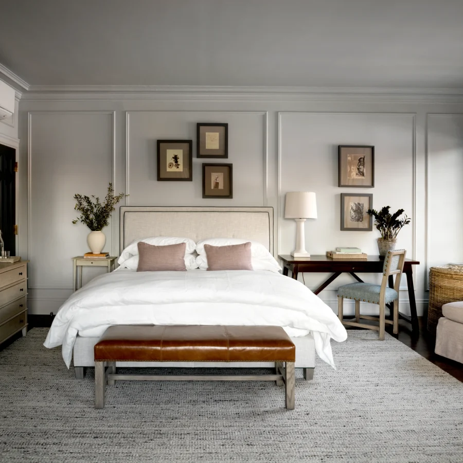 Light gray bedroom with paneled walls, white bed, and framed art above; brown leather bench at foot of bed.