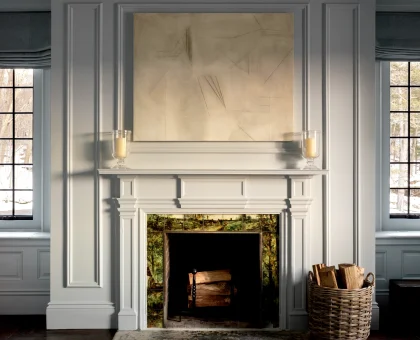 Grey paneled fireplace with an abstract beige painting above, two glass candle holders on the mantel, and a wicker basket of firewood beside a windowed wall.