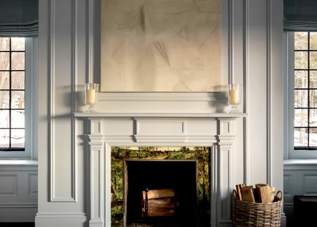 Grey paneled fireplace with an abstract beige painting above, two glass candle holders on the mantel, and a wicker basket of firewood beside a windowed wall.