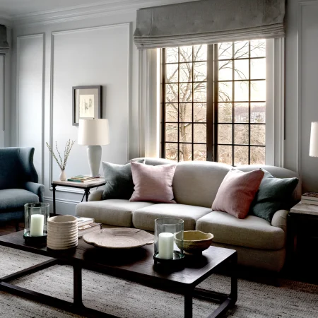 Living room interior with large grid windows, gray walls, and a gray sofa with pink and gray cushions. A blue chair sits to the left, and a wooden coffee table with candles is in the foreground.