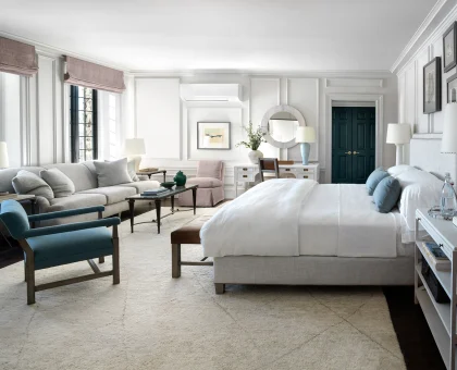 Bright, elegant bedroom with white paneled walls, a large bed, seating area, and teal doors in the distance