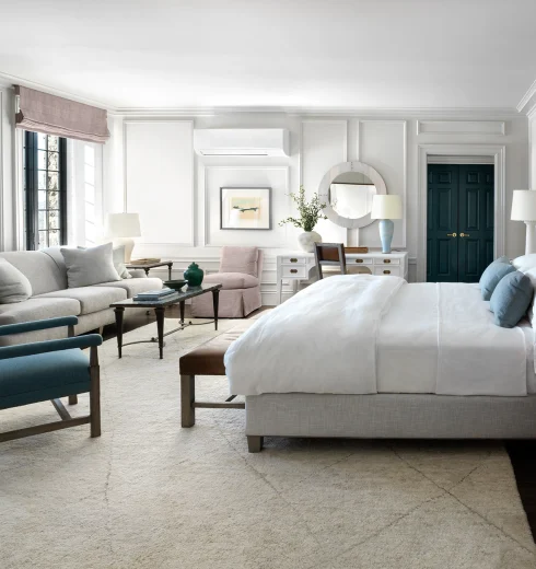 Bright, elegant bedroom with white paneled walls, a large bed, seating area, and teal doors in the distance