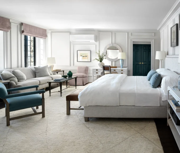 Bright, elegant bedroom with white paneled walls, a large bed, seating area, and teal doors in the distance