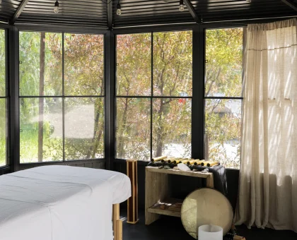 Spa treatment room with two massage tables covered in white sheets, large windows showing trees, and a black vaulted ceiling with hanging light bulbs.