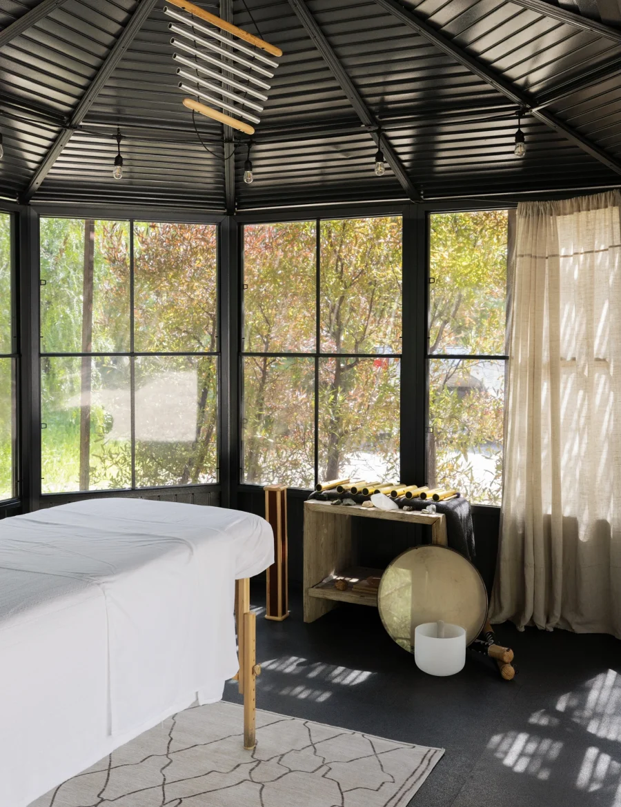Spa treatment room with two massage tables covered in white sheets, large windows showing trees, and a black vaulted ceiling with hanging light bulbs.