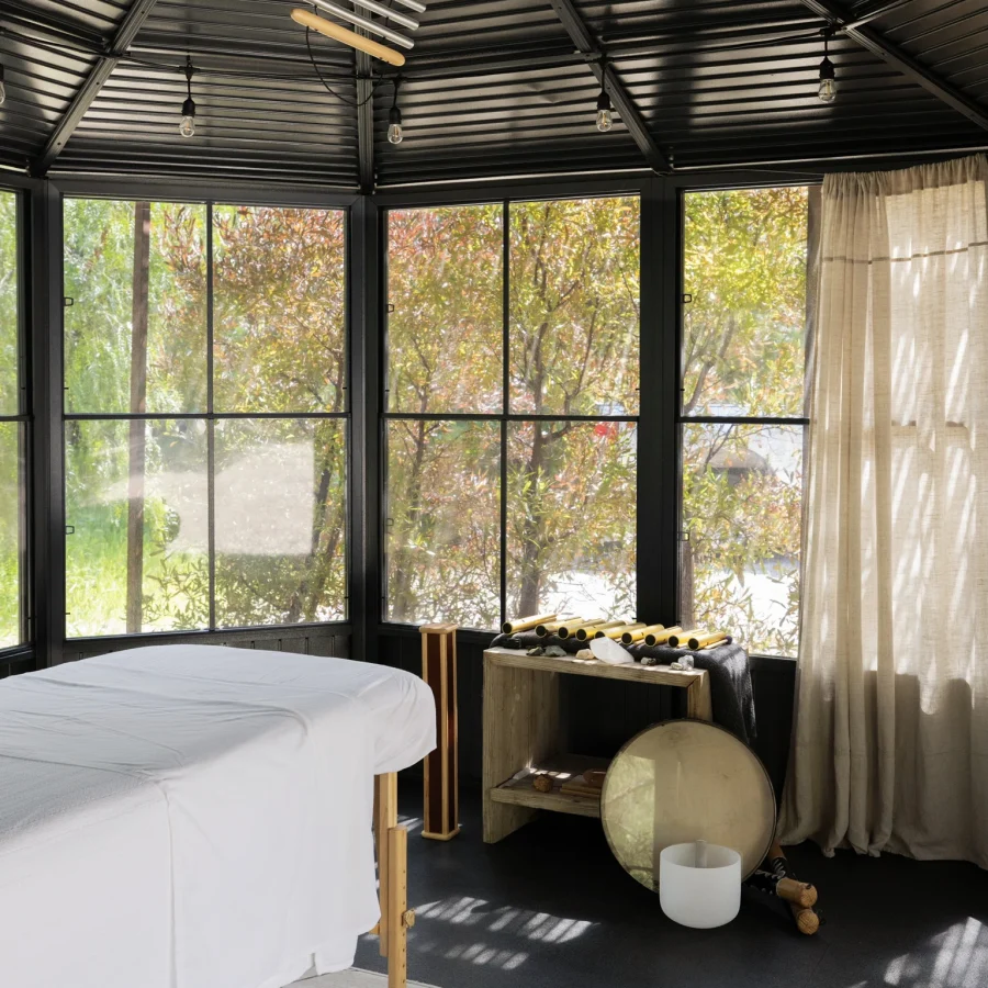 Spa treatment room with two massage tables covered in white sheets, large windows showing trees, and a black vaulted ceiling with hanging light bulbs.