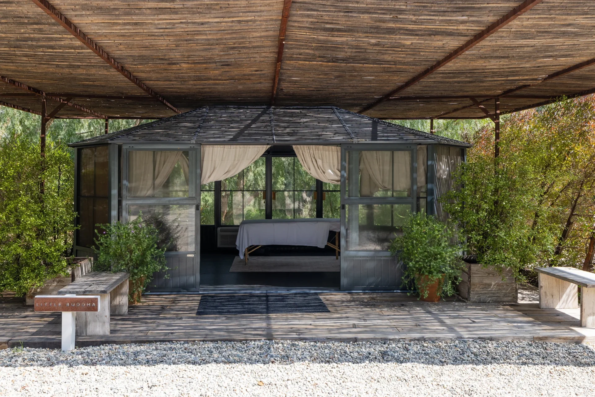A glass-walled bungalow with sheer curtains under a large bamboo pergola, surrounded by greenery and gravel path. Sign reads 'Little Buddha' at left.
