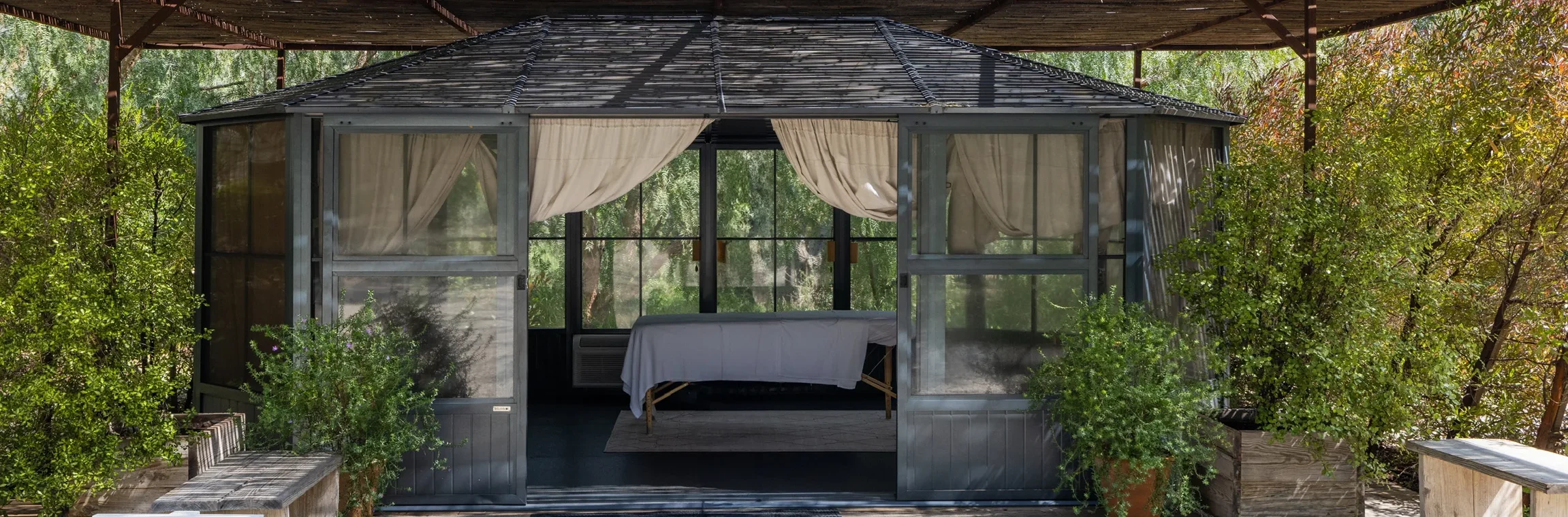 A glass-walled bungalow with sheer curtains under a large bamboo pergola, surrounded by greenery and gravel path. Sign reads 'Little Buddha' at left.