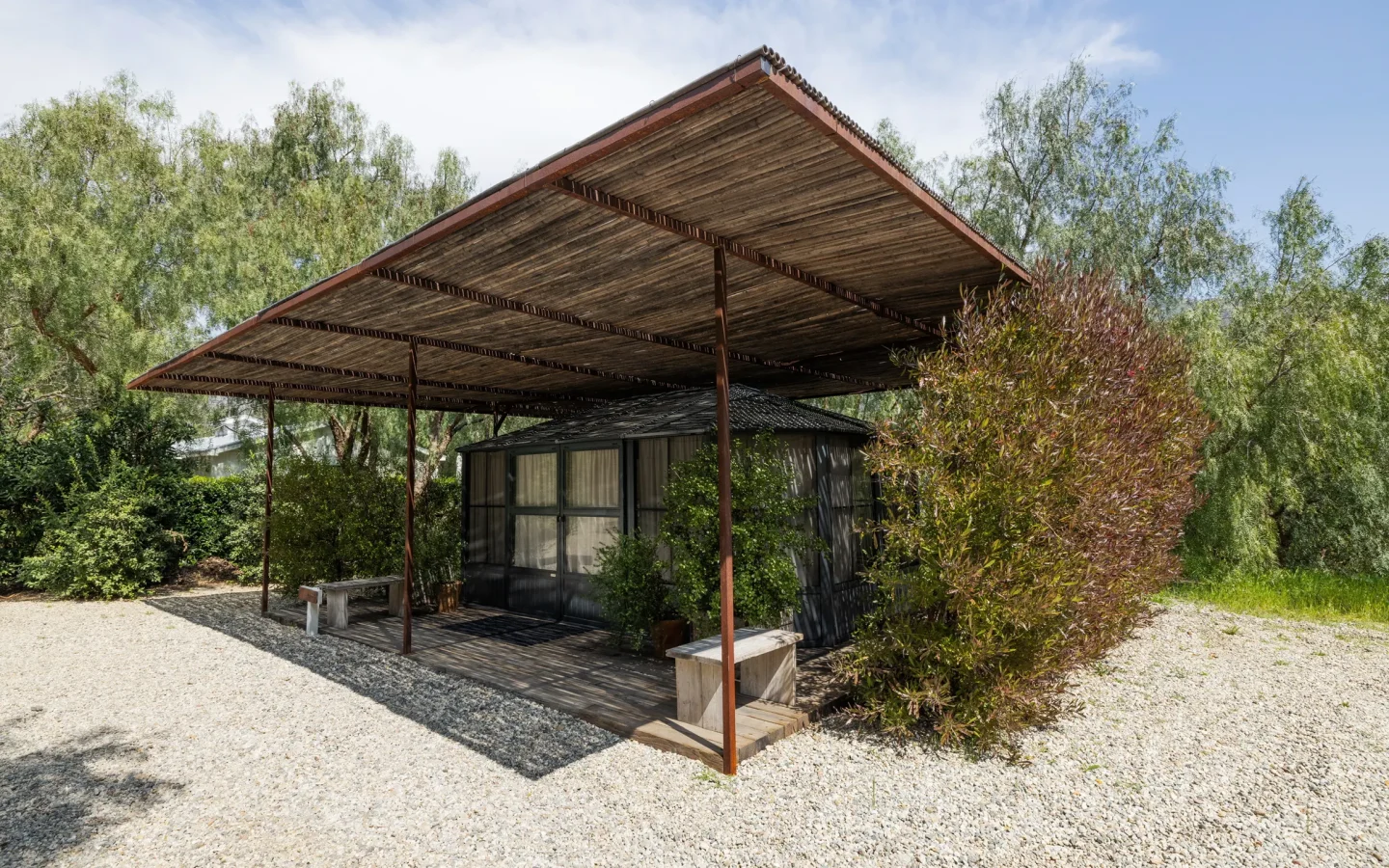 Wooden pergola over a small glass-walled house, surrounded by trees and shrubs on a gravel lot at sunny day.