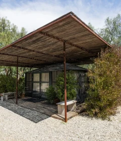 Wooden pergola over a small glass-walled house, surrounded by trees and shrubs on a gravel lot at sunny day.