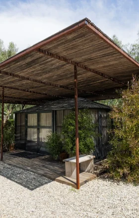 Wooden pergola over a small glass-walled house, surrounded by trees and shrubs on a gravel lot at sunny day.