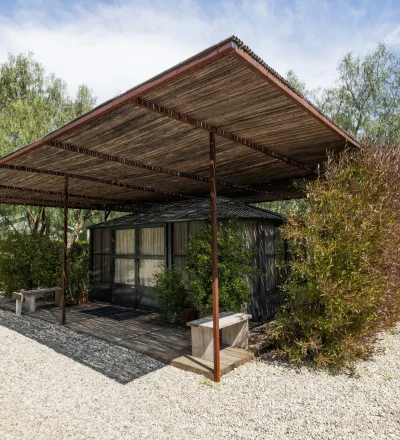 Wooden pergola over a small glass-walled house, surrounded by trees and shrubs on a gravel lot at sunny day.