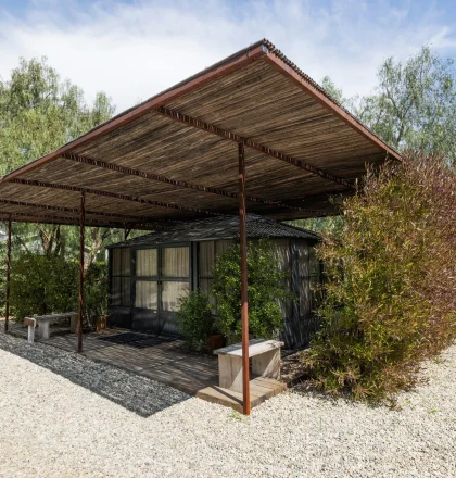 Wooden pergola over a small glass-walled house, surrounded by trees and shrubs on a gravel lot at sunny day.