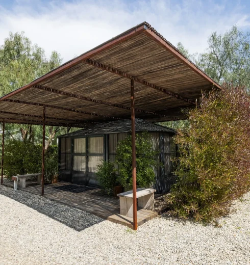 Wooden pergola over a small glass-walled house, surrounded by trees and shrubs on a gravel lot at sunny day.