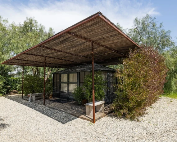Wooden pergola over a small glass-walled house, surrounded by trees and shrubs on a gravel lot at sunny day.