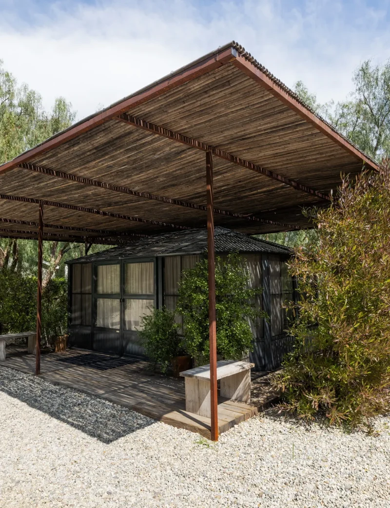 Wooden pergola over a small glass-walled house, surrounded by trees and shrubs on a gravel lot at sunny day.