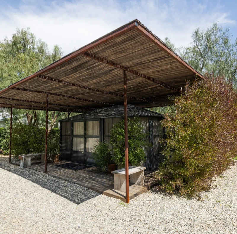 Wooden pergola over a small glass-walled house, surrounded by trees and shrubs on a gravel lot at sunny day.