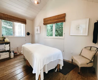 A cozy treatment room with a white-covered massage table, wooden floors, and a pendant light, window blinds, and framed art on the wall