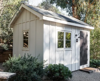 White garden shed with a metal roof, surrounded by trees and a gravel yard.